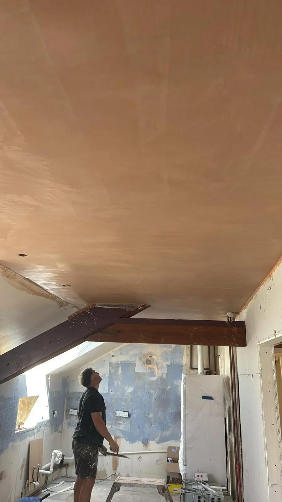 A tradesman holding a trowel looking up at a newly plastered ceiling in a room undergoing construction work